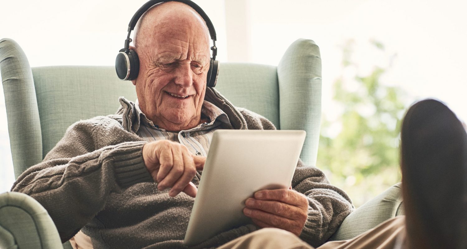 Happy elderly man relaxing on a chair at home and listening to music on digital tablet with headphones